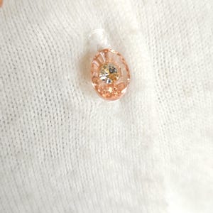 Puede incluir: Primer plano de un c&aacute;rdigan blanco de punto con un bot&oacute;n rosa y adornos de flores rosas.