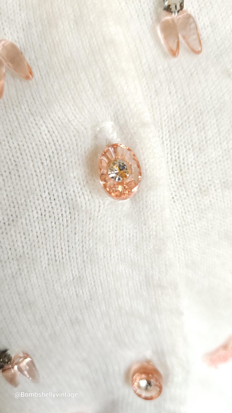 Puede incluir: Primer plano de un c&aacute;rdigan blanco de punto con un bot&oacute;n rosa y adornos de flores rosas.