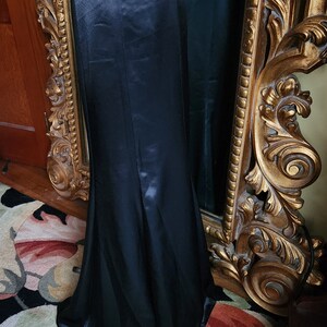 May include: A long, black satin skirt with a high waist and a slight fishtail hem. The skirt is displayed on a mannequin next to a gold ornate mirror. The fabric has a subtle sheen.