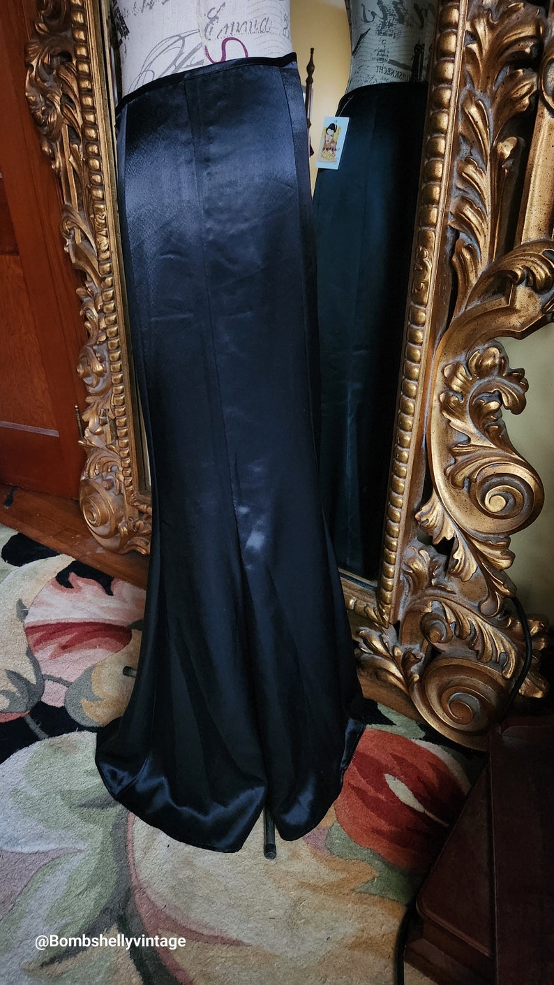 May include: A long, black satin skirt with a high waist and a slight fishtail hem. The skirt is displayed on a mannequin next to a gold ornate mirror. The fabric has a subtle sheen.