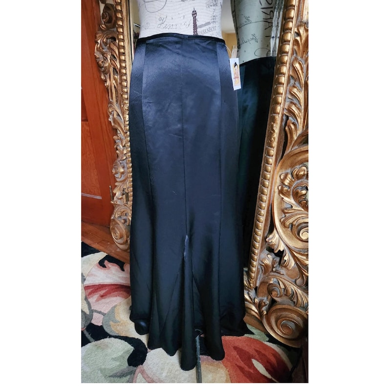May include: A long, black satin skirt with a high waist and a slight flare at the bottom. The skirt has a slit in the front, and is displayed on a mannequin. The skirt is a formal wear item.