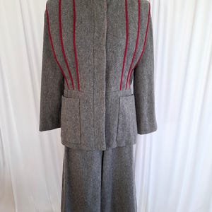 May include: A grey wool-blend two-piece suit consisting of a long-sleeved jacket and a matching skirt. The jacket features a high collar, two front pockets, and vertical red piping accents that extend from the shoulders to the waistline.