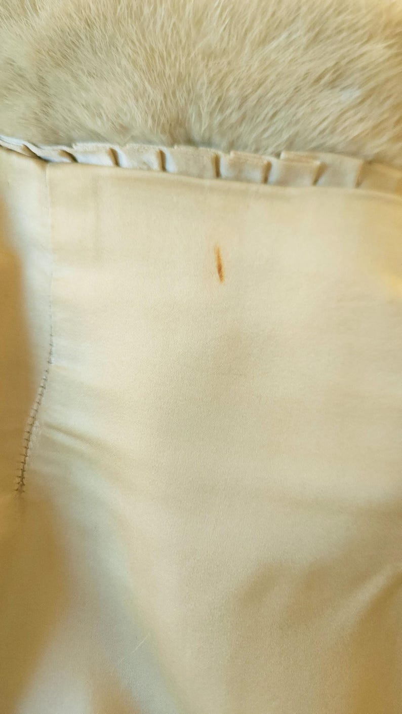 May include: Close-up of a cream-coloured fur coat lining. The lining is a light beige fabric with a small brown stain. The fur is a soft, fluffy texture.