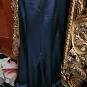 May include: A long, black satin skirt with a high waist and a slight flare at the hem. The skirt is displayed on a mannequin, with a zipper closure visible at the back. The fabric has a subtle sheen, reflecting light.