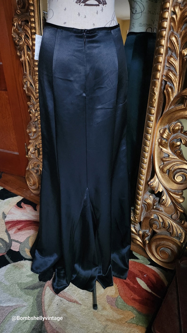 May include: A long, black satin skirt with a high waist and a slight flare at the hem. The skirt is displayed on a mannequin, with a zipper closure visible at the back. The fabric has a subtle sheen, reflecting light.