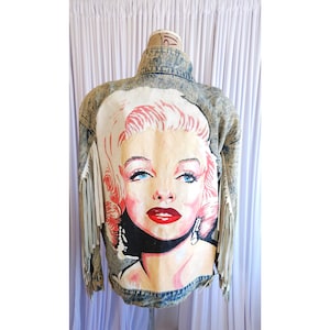 May include: An acid-wash denim jacket featuring a large, hand-painted portrait of a blonde woman with blue eyes and red lips on the back panel. The jacket includes long, light-colored fringe detailing along the sleeves and a classic collar.