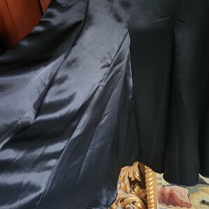 May include: A close-up of a black dress with a flowing skirt. The dress has a satin-like fabric that drapes elegantly. The hemline is visible, and the dress appears to be vintage or high-end. The dress is set against a wooden background.
