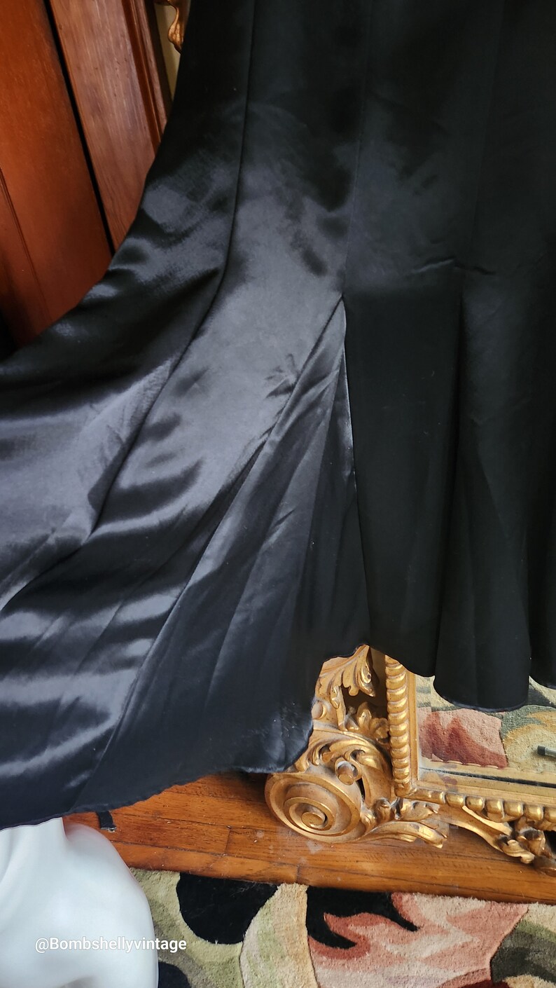 May include: A close-up of a black dress with a flowing skirt. The dress has a satin-like fabric that drapes elegantly. The hemline is visible, and the dress appears to be vintage or high-end. The dress is set against a wooden background.