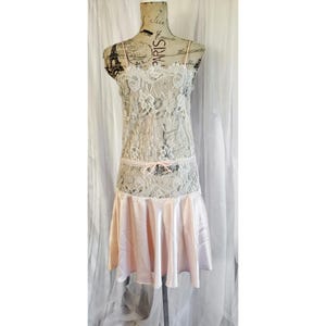 Könnte beinhalten: Vintage Nachthemd auf einer Schaufensterpuppe. Das Kleid hat ein weißes Spitzenoberteil mit Spaghettiträgern und einen hellrosa Satinstoffrock. Eine zarte rosa Schleife verziert die Taille. Das Kleid wird vor weißem Hintergrund präsentiert.