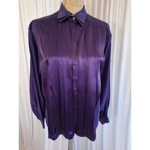 May include: A long-sleeved button-down shirt in a deep purple satin fabric. The shirt features a pointed collar, a front button placket, and vertical pleats running down the front panel. The sleeves are long with buttoned cuffs at the wrists.