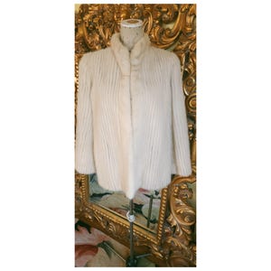 May include: An ivory-coloured vintage coat with a high collar and vertical ribbed texture. The coat has long sleeves and a soft, plush appearance. The coat is displayed on a mannequin.