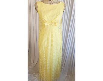 May include: A sleeveless, floor-length, pale yellow lace dress. The dress features a bow detail at the waist and a scoop neckline. The dress is displayed on a mannequin against a white backdrop.