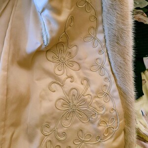 May include: Detail of a vintage coat's interior. The cream-coloured satin lining is adorned with embroidered floral patterns in a matching thread. The edge is trimmed with light brown fur.