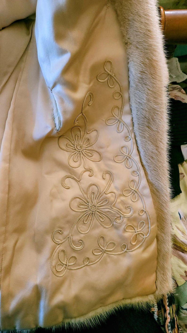 May include: Detail of a vintage coat's interior. The cream-coloured satin lining is adorned with embroidered floral patterns in a matching thread. The edge is trimmed with light brown fur.