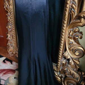 May include: A long, black satin skirt with a slight fishtail hem, displayed on a mannequin. The skirt has a high waist and a subtle sheen. A vintage-style gold mirror is in the background.