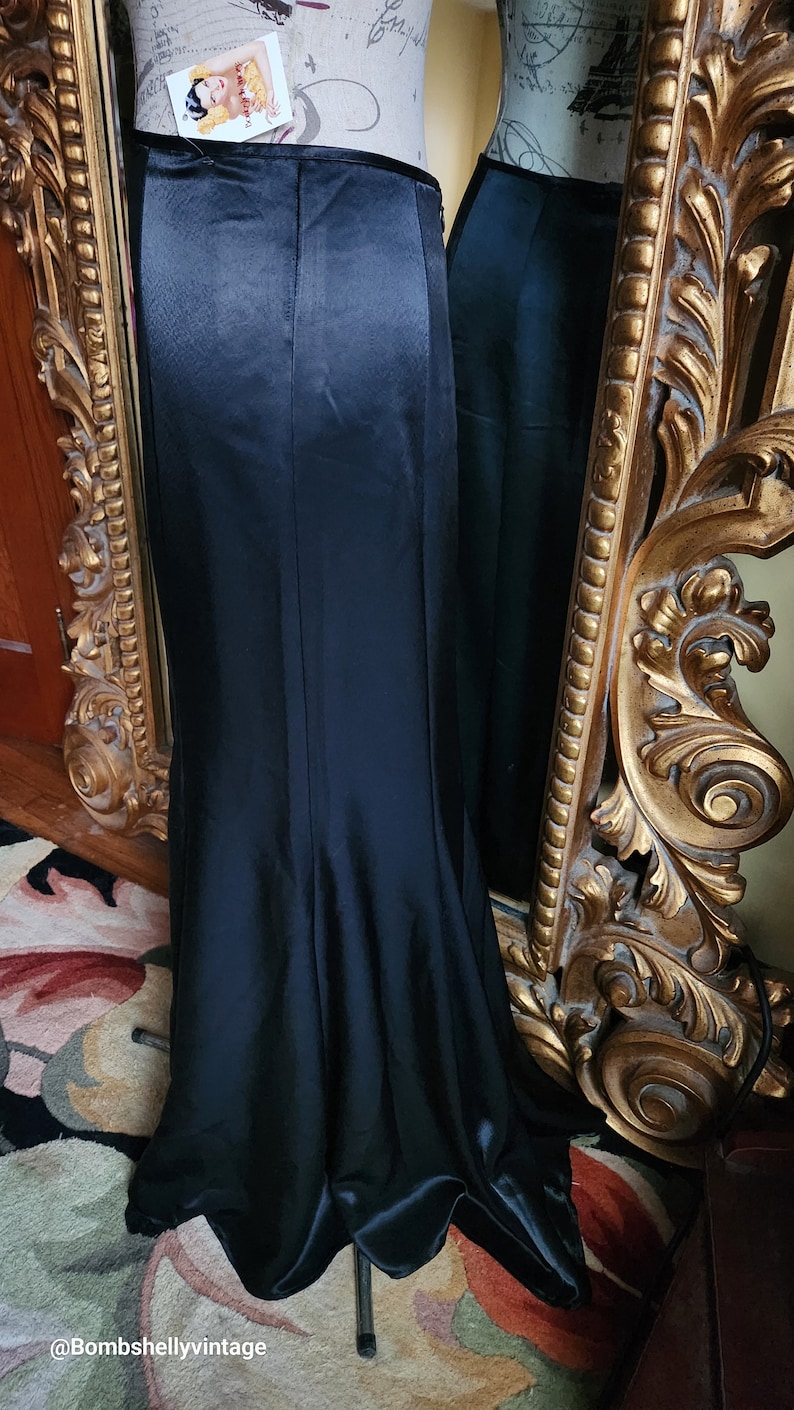 May include: A long, black satin skirt with a slight fishtail hem, displayed on a mannequin. The skirt has a high waist and a subtle sheen. A vintage-style gold mirror is in the background.
