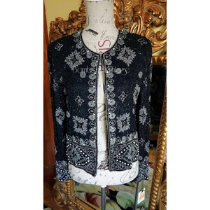 Könnte beinhalten: Schwarze Perlenjacke mit silbernen und weißen floralen und geometrischen Mustern. Die Jacke hat einen runden Ausschnitt und lange Ärmel. Die Jacke ist vorne offen und trägt den Text "Bombs Vintage".