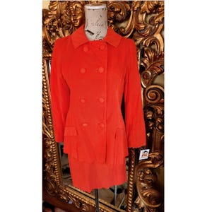 Puede incluir: Un traje de color naranja brillante de doble botonadura con falda y chaqueta. El traje está en un maniquí delante de un espejo dorado adornado.