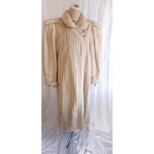 May include: A long, light beige fur coat with a large collar and long sleeves. The coat is displayed on a mannequin and features a circular clasp closure. The fur appears soft and luxurious, with a vertical pattern.