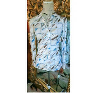 Puede incluir: Camisa de manga larga con un estampado geométrico en blanco, marrón y azul. La camisa tiene cuello puntiagudo y se exhibe en un maniquí. El diseño presenta cuadrados y líneas diagonales.