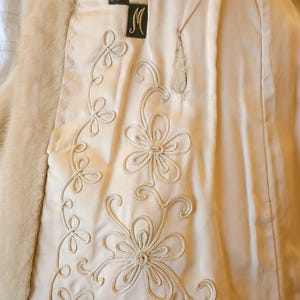 May include: Close-up of a cream-coloured coat interior with embroidered floral detailing. The coat features a faux fur trim and decorative stitching. The design includes the letters "W" and "M" on black patches, and a purple emblem.