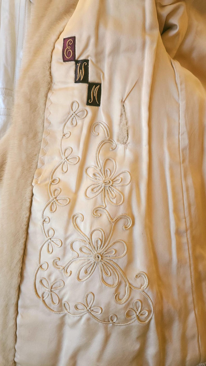 May include: Close-up of a cream-coloured coat interior with embroidered floral detailing. The coat features a faux fur trim and decorative stitching. The design includes the letters "W" and "M" on black patches, and a purple emblem.