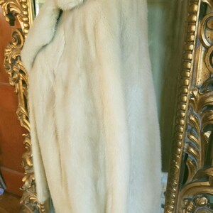 May include: A cream-coloured fur coat with a wide collar, displayed on a mannequin. The coat has a soft, plush texture and a slightly curved hem. The background includes a gold-framed mirror.