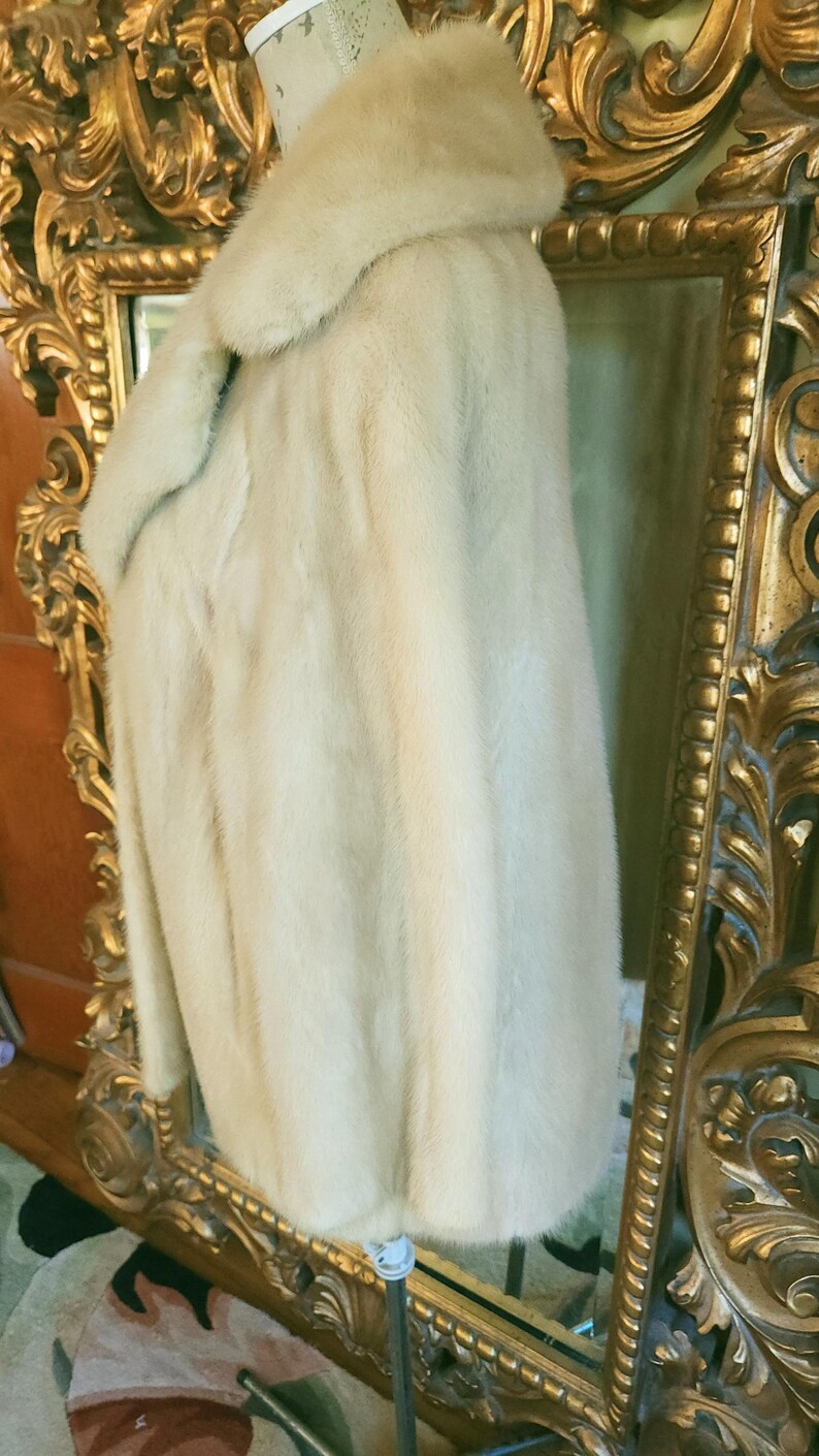 May include: A cream-coloured fur coat with a wide collar, displayed on a mannequin. The coat has a soft, plush texture and a slightly curved hem. The background includes a gold-framed mirror.