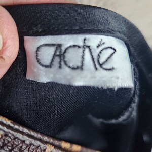 May include: Close-up of a black fabric with a white label that reads "Cache" in a stylized font. The label is sewn onto the fabric. A brown and tan patterned strap is visible.