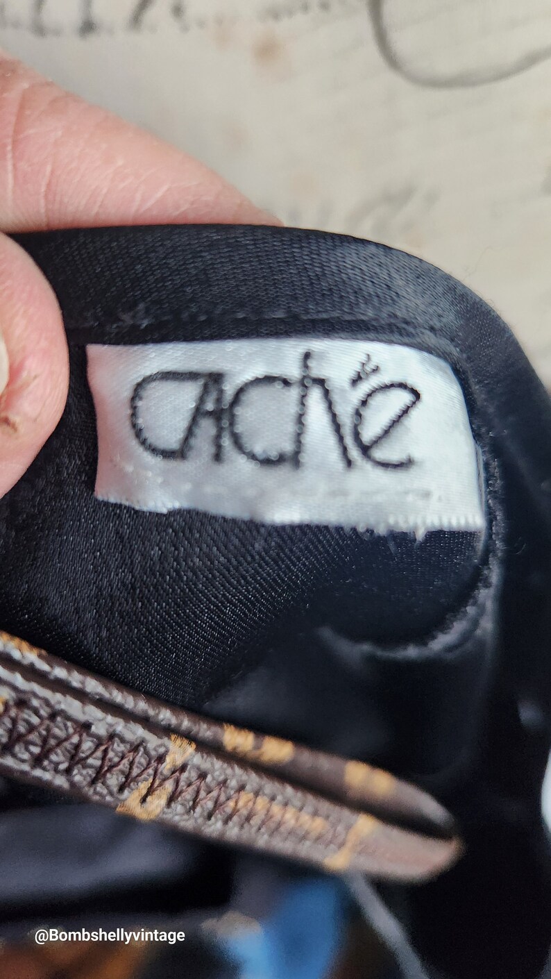 May include: Close-up of a black fabric with a white label that reads "Cache" in a stylized font. The label is sewn onto the fabric. A brown and tan patterned strap is visible.