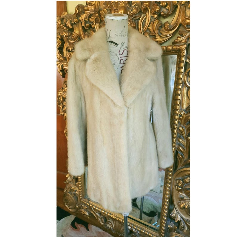 May include: A pale beige fur coat with a wide collar and long sleeves. The coat is displayed on a mannequin in front of a gold-framed mirror. The coat has a classic, elegant design.