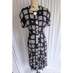 Puede incluir: Un vestido de largo midi con un patrón de cuadrícula en blanco y negro y un diseño cuadrado texturizado. La prenda tiene mangas cortas, cuello alto y una tapeta de botones frontal con botones blancos grandes y redondos desde el cuello a la cintura.