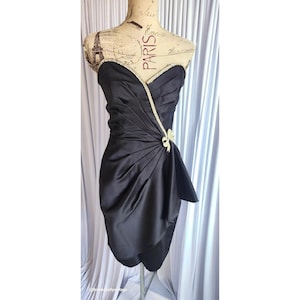 Puede incluir: Vestido de cóctel negro sin tirantes con escote corazón. El vestido presenta un corpiño drapeado con un lazo en la cintura y un ribete de cuentas a lo largo del escote. El vestido se exhibe en un maniquí.