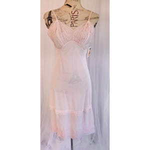 Könnte beinhalten: Ein hellrosa Unterkleid mit zarten Spitzendetails. Das Kleid hat Spaghettiträger und einen V-Ausschnitt mit Spitzensaum. Das Oberteil und der Saum sind mit Spitze verziert, was einen Vintage-Look erzeugt. Das Kleid wird auf einer Schaufensterpuppe präsentiert.