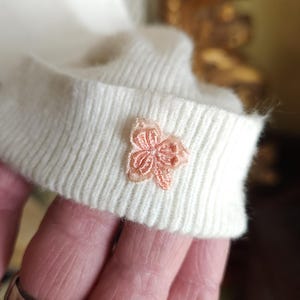 Puede incluir: Un su&eacute;ter de punto blanco con una flor rosa bordada en el pu&ntilde;o.