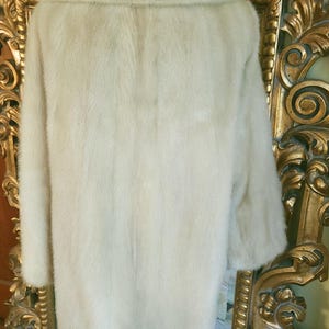 May include: A pale fur coat with a broad collar and long sleeves. The coat is displayed on a mannequin in front of a gold-framed mirror. The fur appears soft and plush, with a subtle sheen.
