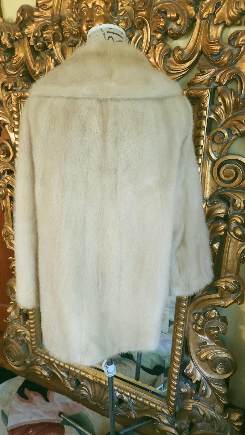 May include: A pale fur coat with a broad collar and long sleeves. The coat is displayed on a mannequin in front of a gold-framed mirror. The fur appears soft and plush, with a subtle sheen.