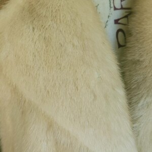 May include: Close-up of a cream-coloured fur coat. The fur appears soft and plush, with a slight sheen. The collar is visible, and the word "PARIS" is partially visible on the lining.