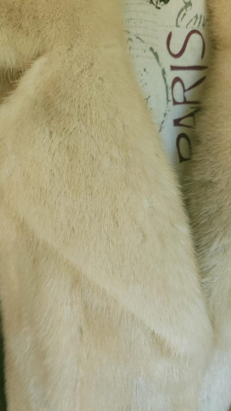 May include: Close-up of a cream-coloured fur coat. The fur appears soft and plush, with a slight sheen. The collar is visible, and the word "PARIS" is partially visible on the lining.