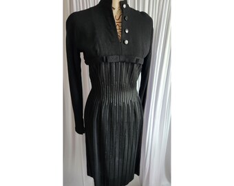 Könnte beinhalten: Ein schwarzes Vintage-Kleid mit langen Ärmeln und einem hohen Kragen. Das Kleid hat ein figurbetontes Oberteil mit vertikalen Rippendetails und dekorativen Knöpfen. Das Kleid wird auf einer Schaufensterpuppe präsentiert.