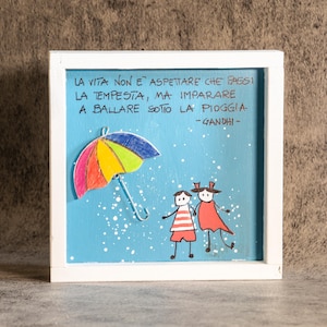 Könnte beinhalten: Ein gerahmtes Kunstwerk mit hellblauem Hintergrund zeigt einen bunten Regenschirm und zwei Strichmännchen im Regen. Der italienische Text lautet: "LA VITA NON E ASPETTARE CHE PASSI LA TEMPESTA, MA IMPARARE A BALLARE SOTTO LA PIOGGIA - GANDHI".