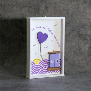 Può includere: Una targa da parete rettangolare in legno bianco con un palloncino a forma di cuore viola legato con uno spago a una piccola casa viola. Il testo "La' dove sei felice sei a casa" è scritto in viola. Sotto ci sono onde viola e un sole giallo.