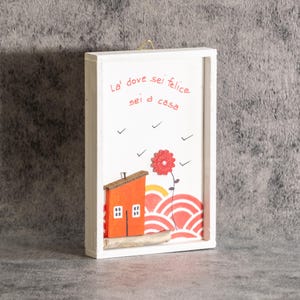 Può includere: Una cornice rettangolare in legno bianco contenente un'opera da parete con una casa arancione, un fiore rosso e motivi a onde rossi. Il testo "La' dove sei felice sei a casa" è stampato in rosso. Un piccolo pezzo di legno galleggiante è alla base.