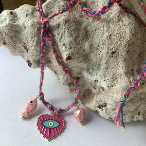 Peut inclure: Un collier en cordon tressé rose et bleu avec un accent doré. Le collier présente un pendentif en forme de cœur rose avec un motif d'œil bleu maléfique, un pendentif en forme de poisson rose à pois blancs et un pendentif en forme de cœur rose.