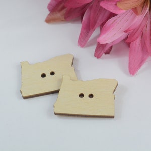 May include: Two wooden buttons shaped like the state of Oregon. Each button has two holes for sewing.