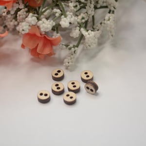 May include: A collection of eight small, round wooden buttons with two holes each. The buttons have a light brown center and a dark brown rim. Peach-colored flowers and white baby's breath are in the background.
