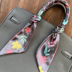 May include: A dark gray handbag with a decorative silk scarf tied to the handle. The scarf features a colorful leaf pattern in shades of red, yellow, and green, with a pink border. The bag is made of textured leather.