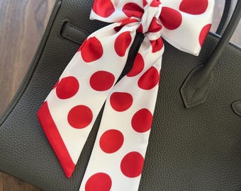 May include: A white scarf with red polka dots tied to a dark grey leather handbag. The scarf has a red border and large red circles on a white background. The handbag has a structured design.