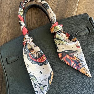May include: A beige silk scarf with a colourful tattoo-inspired design is tied around the handle of a dark green leather handbag.