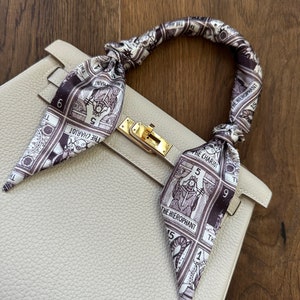 May include: A light gray leather handbag with a gold clasp and a silk scarf tied around the handle. The scarf features a black and white print of tarot cards with the names "The Chariot", "The Hierophant", and "The Hanged Man".